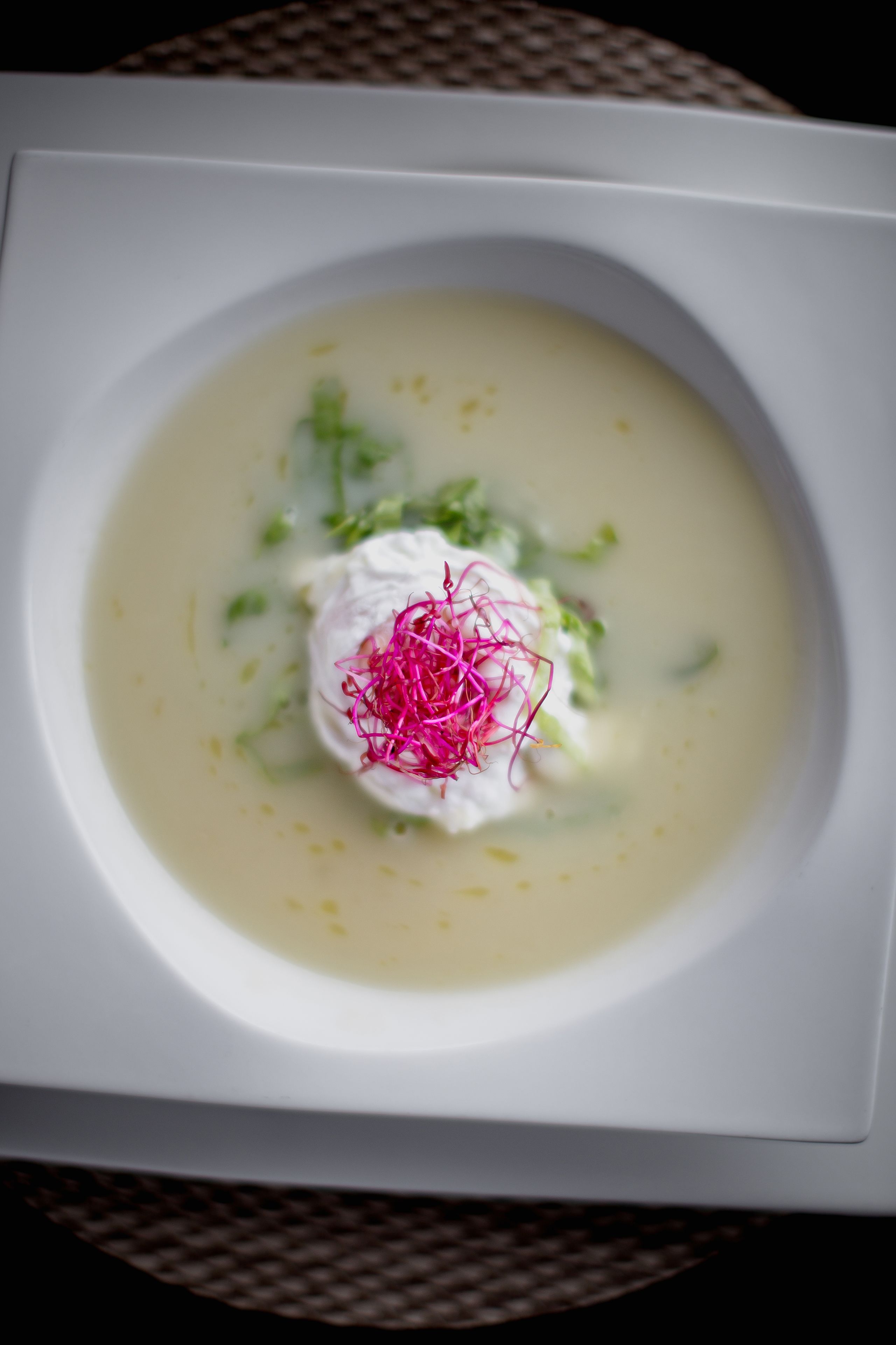A white square plate with a creamy soup, garnished with a dollop of cream and pink edible flowers, and green herbs.