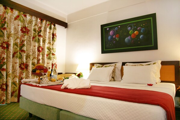 A hotel room with a bed, a fruit bowl, a bottle of water, and a painting on the wall.