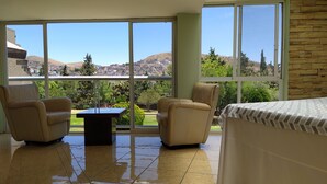 A room with a bed, two chairs, a small table, and large windows offering a view of a hillside town. - Parador (ZACATECAS)