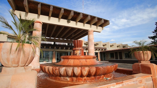 A fountain with a tiered design, surrounded by large planters and a building with a tiled roof in the background. - Parador (ZACATECAS)