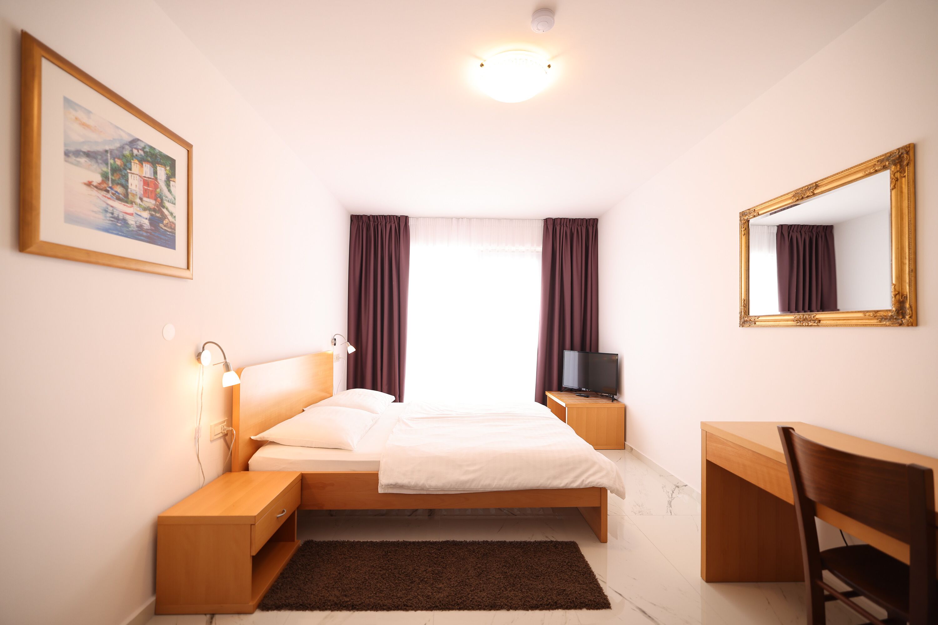 A bedroom with a bed, bedside tables, a desk, a chair, a television, a painting, and a window with curtains.