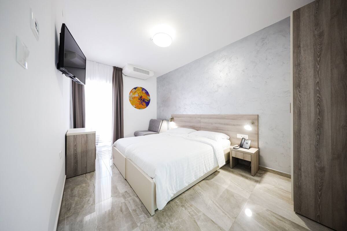 Una habitación de hotel moderna con una cama grande, mesitas de noche, un televisor de pantalla plana en la pared y un amplio ventanal con cortinas.