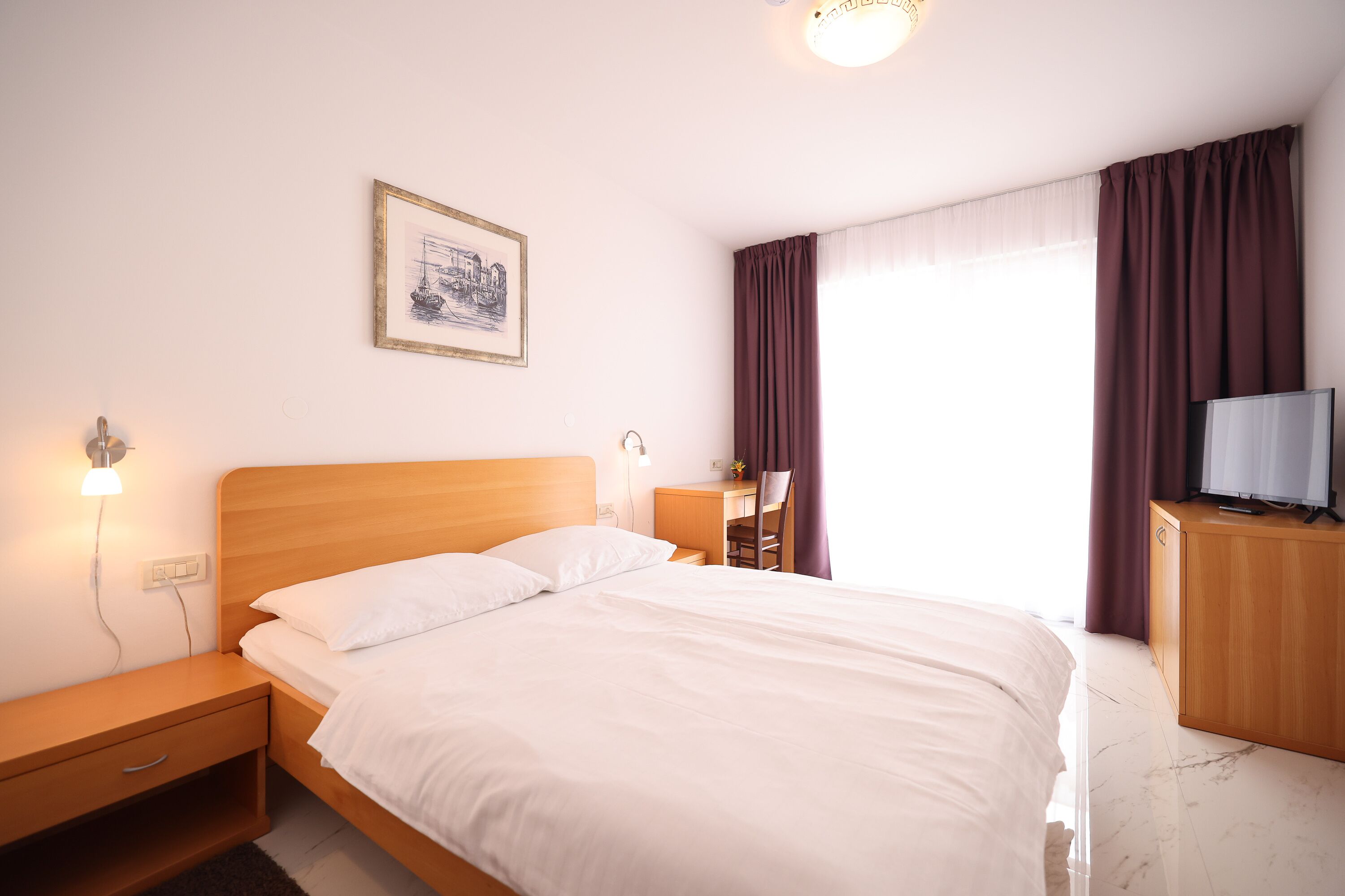 A neatly made bed with white linens, a wooden nightstand with a lamp, a desk with a chair, a television on a wooden cabinet, and a framed picture on the wall.