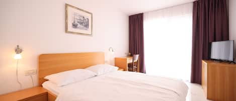 A neatly made bed with white linens, a wooden nightstand with a lamp, a desk with a chair, a television on a wooden cabinet, and a framed picture on the wall.