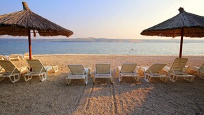 Ein Strand mit sechs Liegestühlen und zwei Reetsonnenschirmen.