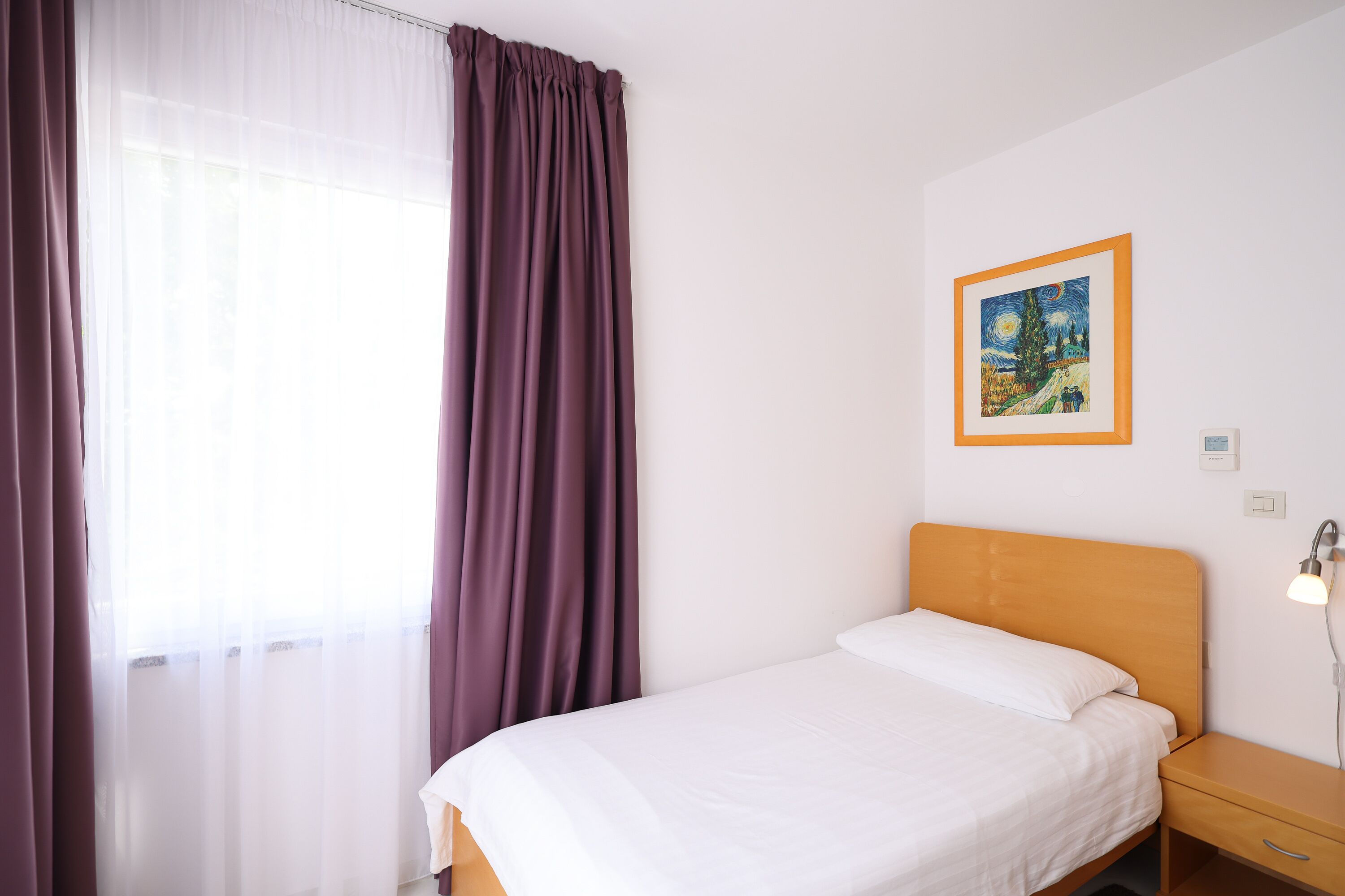 Una cama individual con cabecera de madera, una mesita de noche con lámpara, un cuadro en la pared y cortinas de encaje en la ventana.