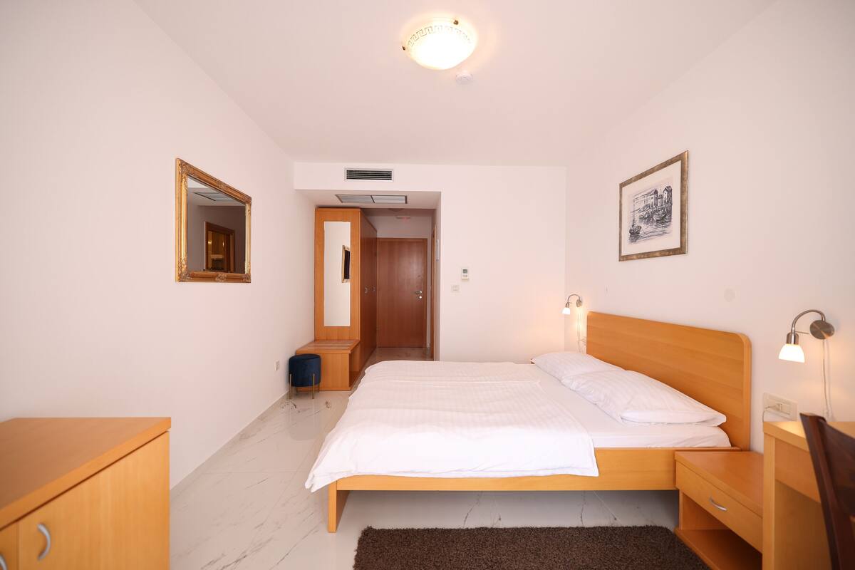 Un dormitorio con cama de madera, mesitas de noche, cómoda, espejo, un cuadro en la pared y una puerta que da a otra habitación.