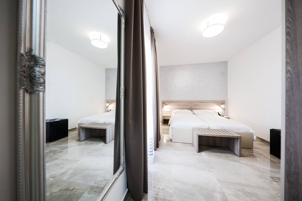 Una habitación de hotel moderna con un espejo grande, dos camas, un banco de madera y un suelo de mármol.