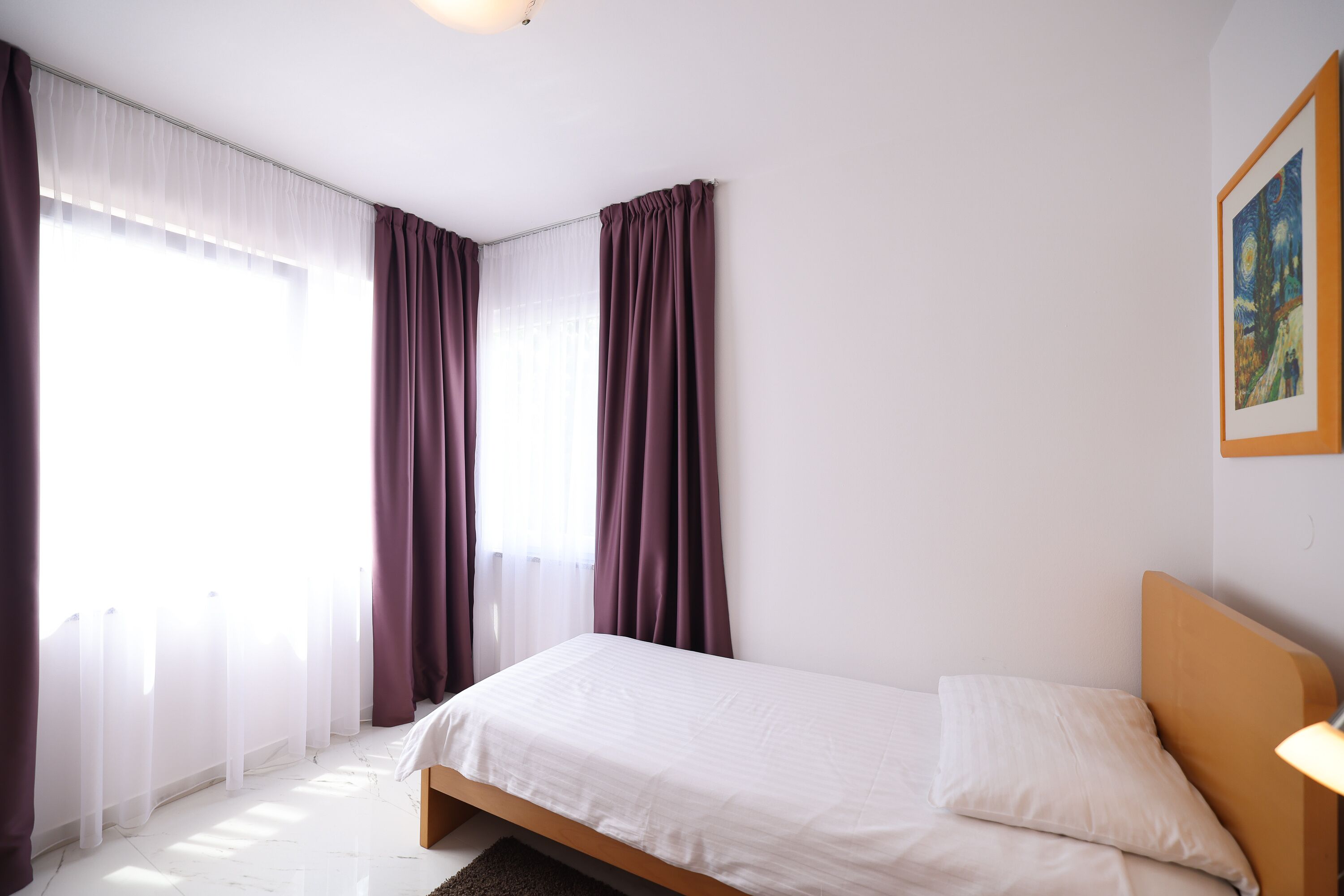 A bedroom with a single bed, white bedding, a wooden headboard, a nightstand with a lamp, and a painting on the wall.