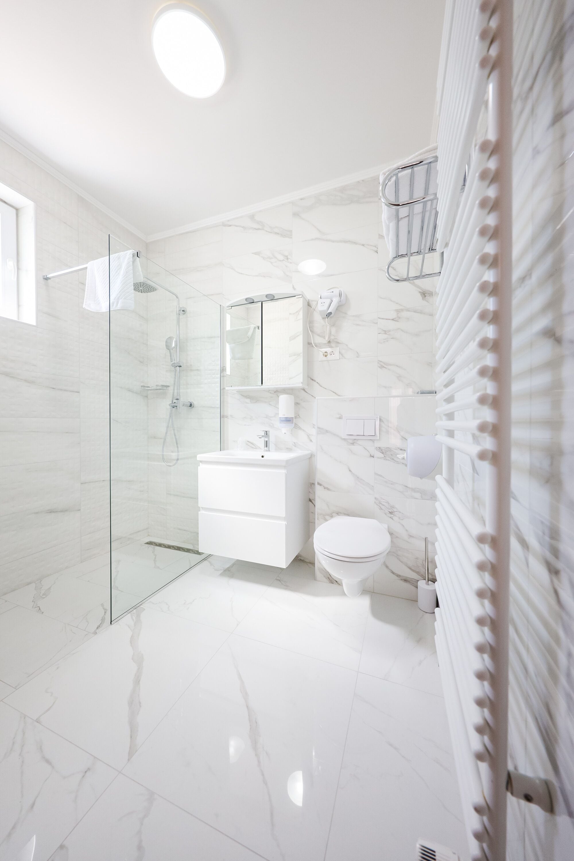 Un baño moderno con ducha de vidrio, azulejos de mármol blanco, tocador blanco y un inodoro.