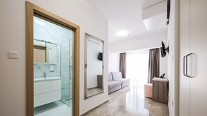 Ein modernes Hotelzimmer mit Glasdusche, einem großen Spiegel, einer Couch, einem Holzkabinett und einem an der Wand montierten Flachbildfernseher.