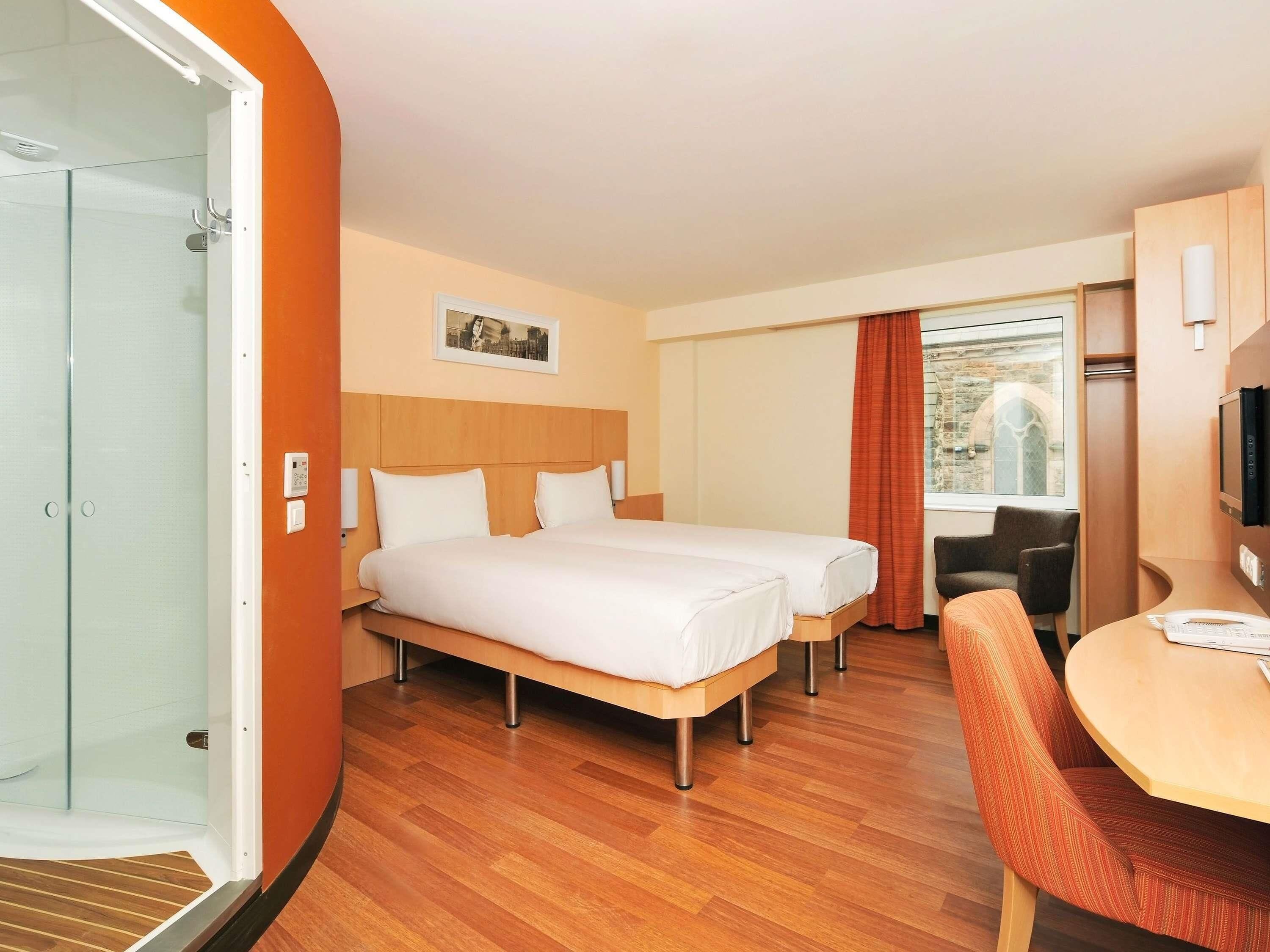 A hotel room with two beds, a desk, a chair, a television, and a window with a view of a building.