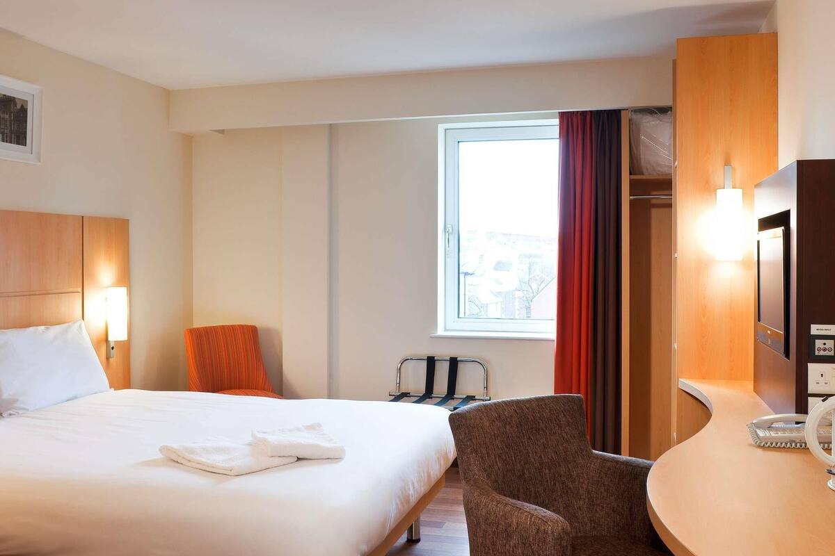 A hotel room with a bed, a chair, a desk, a TV, and a window with curtains.