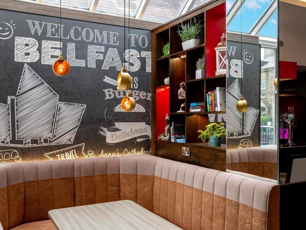 A restaurant with a chalkboard wall featuring 'WELCOME TO BELFAST' and 'ENJOY OUR BURGERS,' a seating area with cushioned booths, and a bookshelf with books and decorative items. - ibis Belfast Queens Quarter (Belfast)