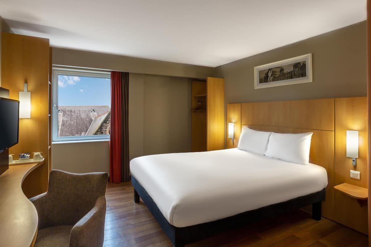 Une chambre dâhĂŽtel avec un grand lit, un bureau avec une chaise, une tĂ©lĂ©vision, une fenĂȘtre avec des rideaux et une vue sur la ville.