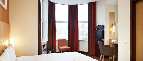 A hotel room with a large bed, a desk with a chair, a television, and a window with curtains.
