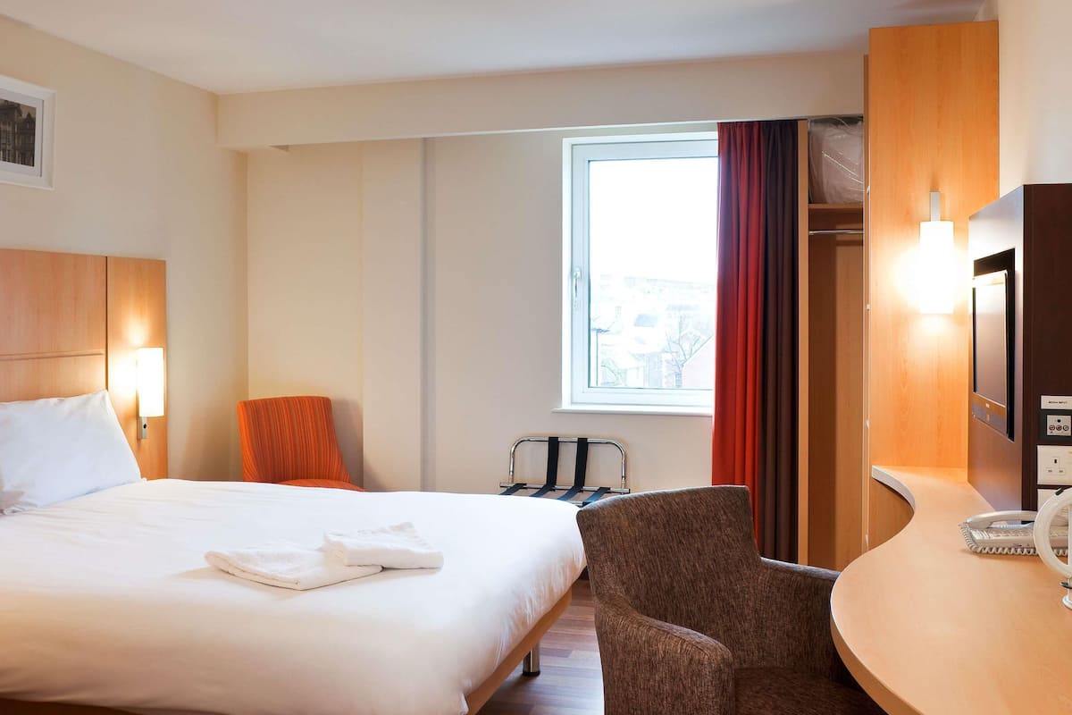 A hotel room with a bed, a chair, a desk, a TV, and a window.