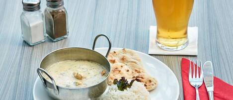 A plate with rice, naan bread, and a metal bowl of curry, a glass of beer, salt and pepper shakers, and cutlery on a wooden table.