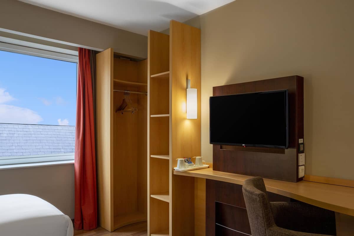 A hotel room with a bed, a desk with a TV, a wardrobe, and a window with curtains.