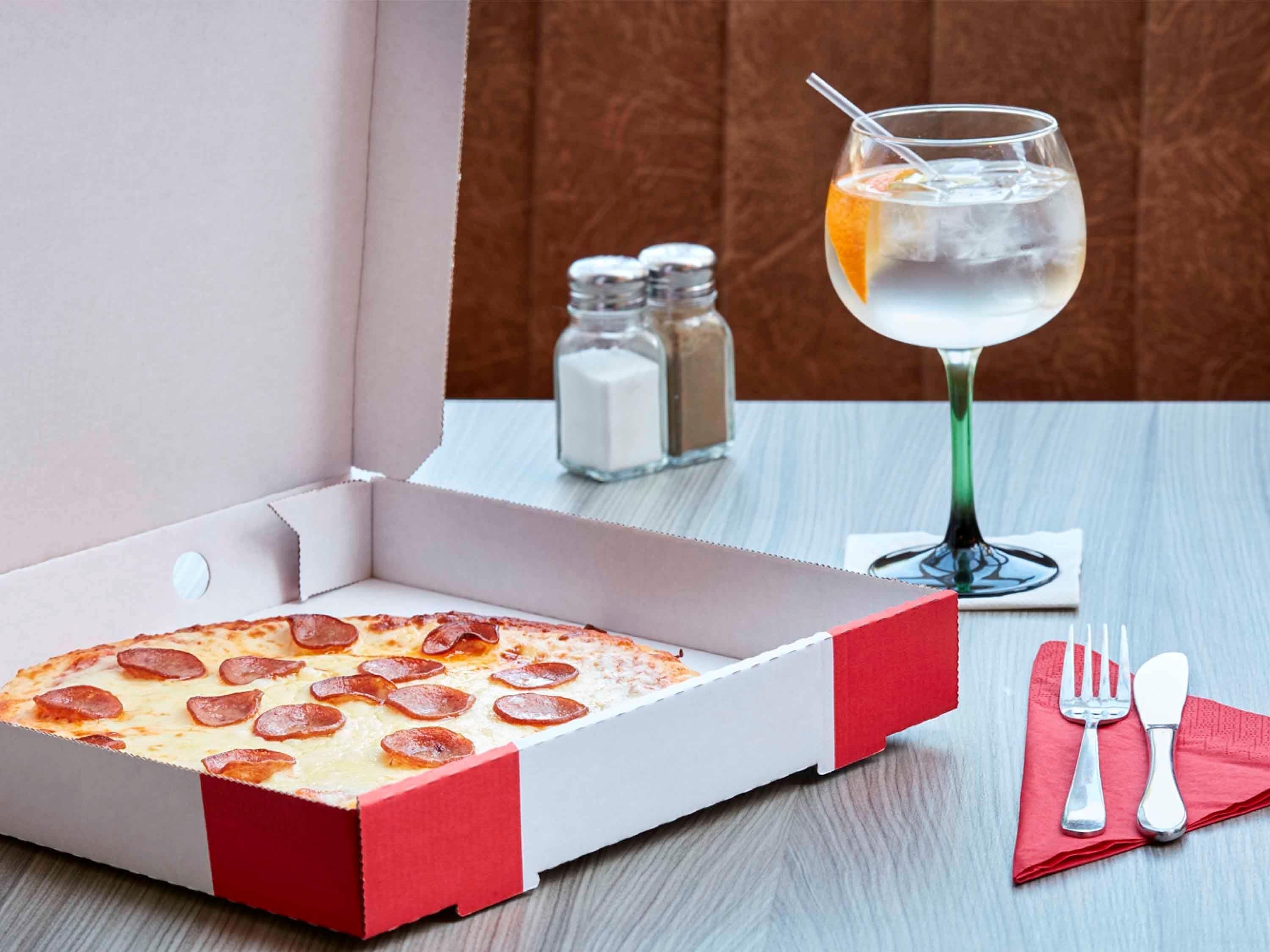 A pizza box with pepperoni slices on a table, a glass of iced drink, salt and pepper shakers, and a napkin with cutlery.