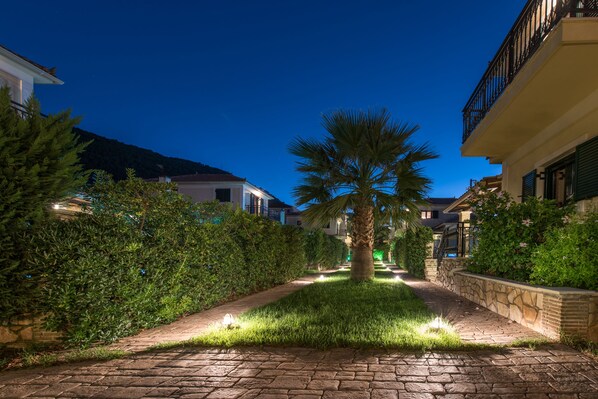 A paved pathway flanked by lush greenery and a palm tree, leading to a building with a balcony.