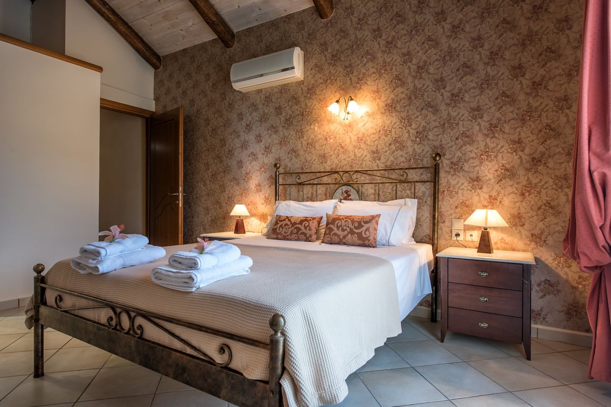 A neatly made bed with pillows and folded towels, a bedside table with a lamp, and a floral wallpapered wall.