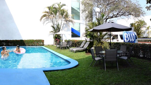 A swimming pool with a blue edge, surrounded by grass and palm trees. There are lounge chairs and a table with umbrellas. - Best Western Global Express (Veracruz)