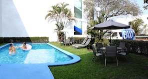 A swimming pool with a blue edge, surrounded by grass and palm trees. There are lounge chairs and a table with umbrellas.