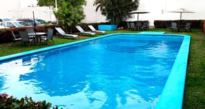 Una piscina rectangular con borde azul, rodeada de césped, sillas y sombrillas.