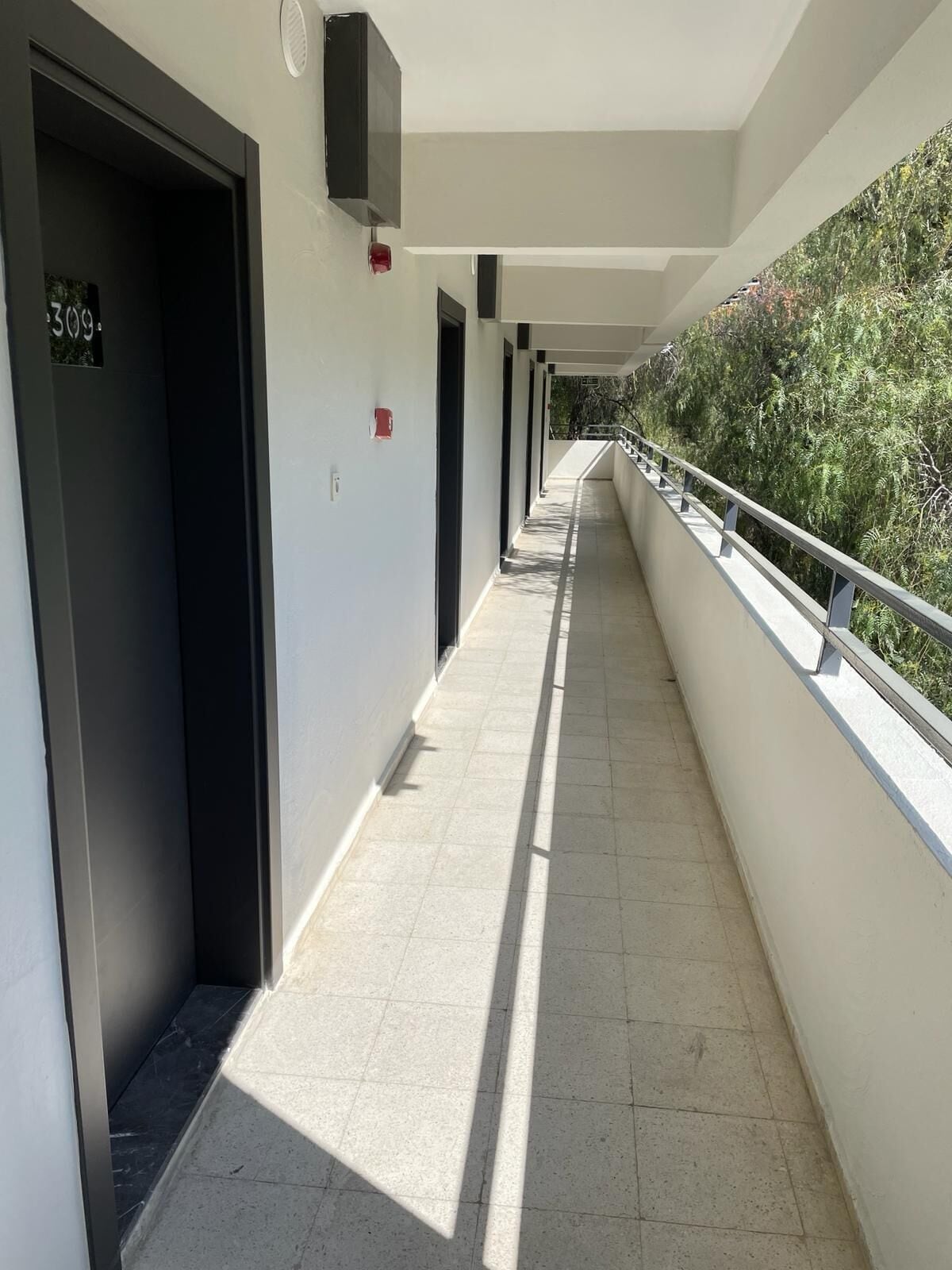 A corridor with doors, a fire alarm, and a railing overlooking a vegetated area.
