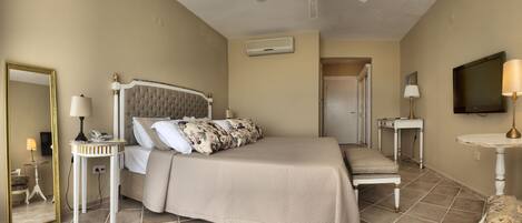 A bedroom with a bed, bedside tables, a mirror, a ceiling fan, a TV, and a door leading to another room.