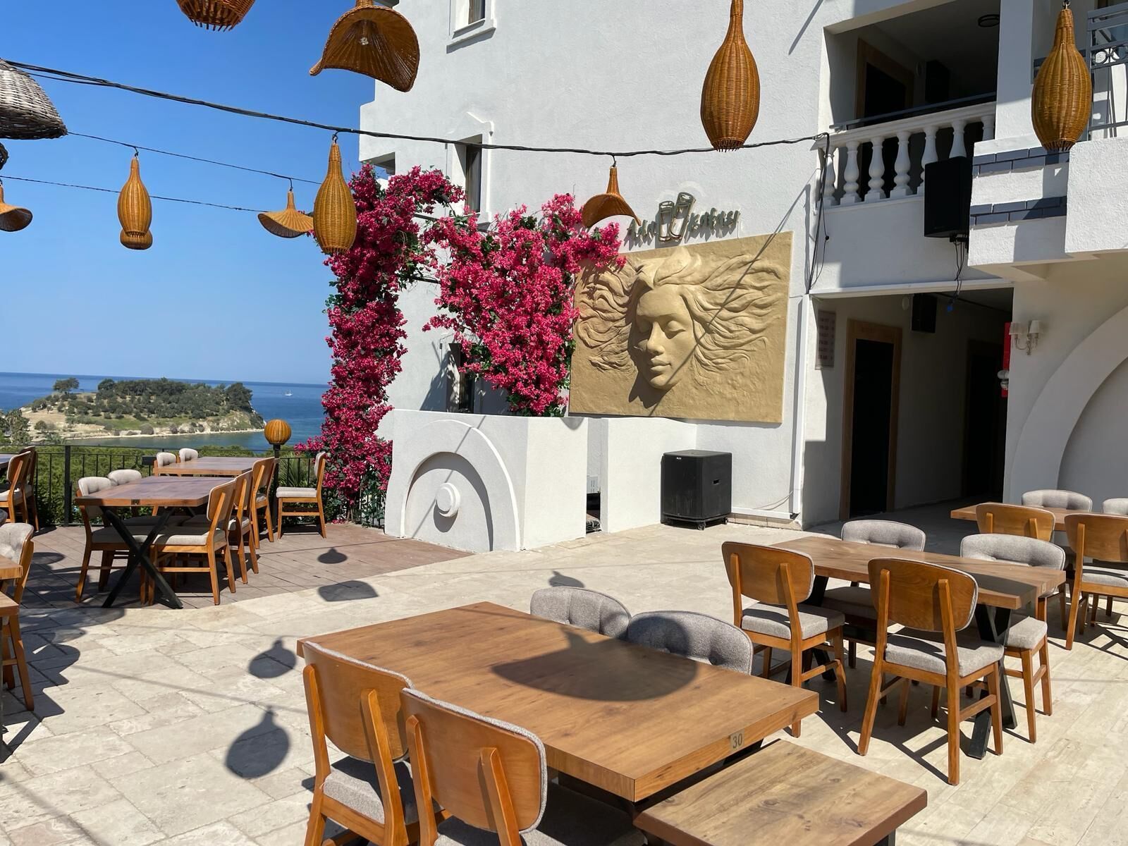An outdoor seating area with wooden tables and chairs, a decorative wall with a woman's face, and a view of the sea.