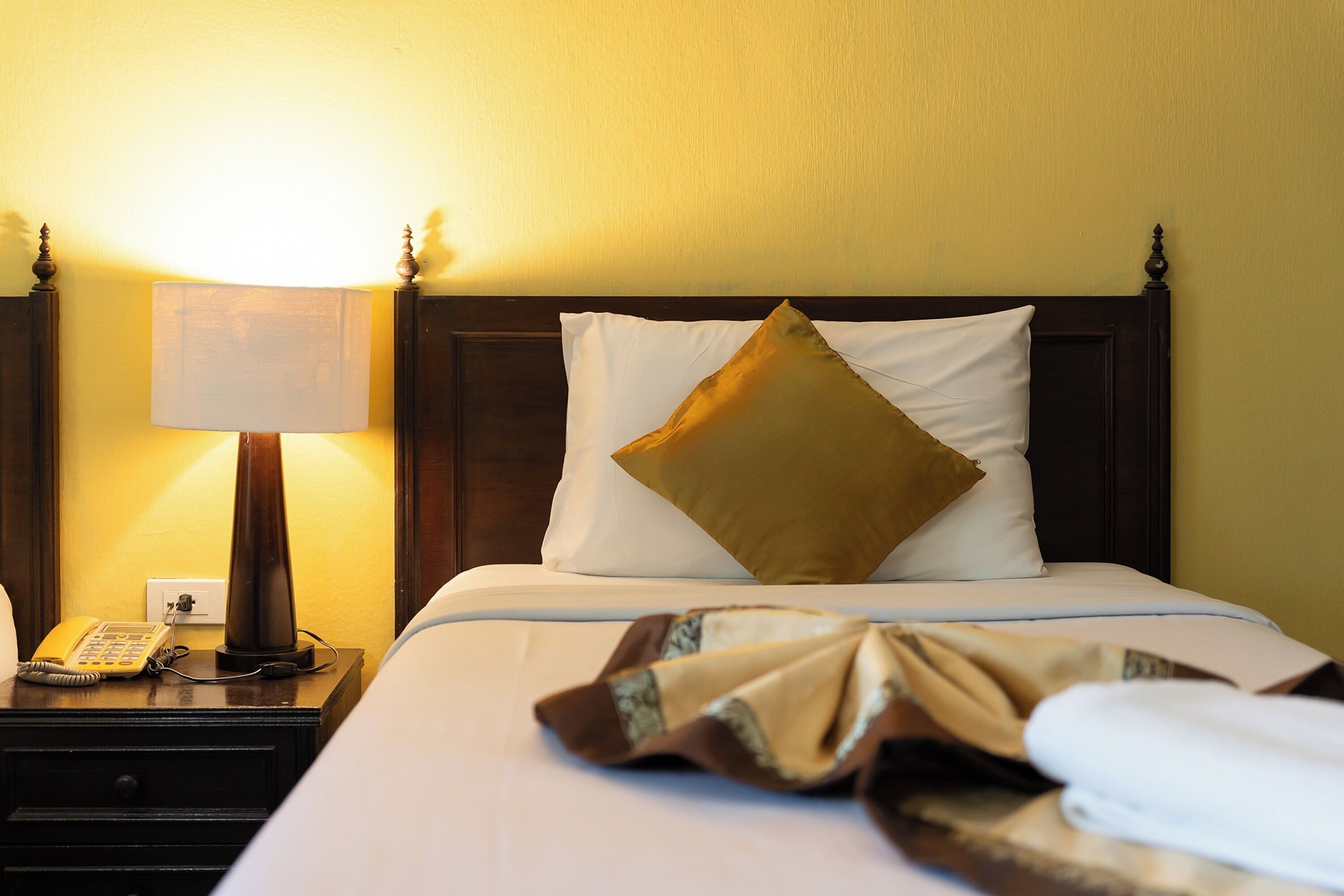 Una cama bien tendida, con almohada blanca y dorada, una lámpara de noche y un teléfono sobre una mesita de madera.