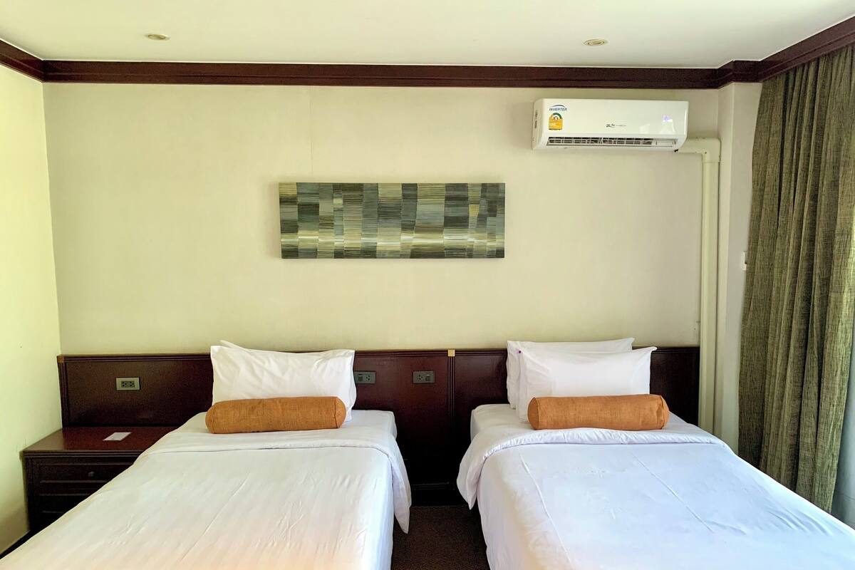 A hotel room with two single beds, a nightstand, a wall-mounted air conditioner, and a painting above the beds.