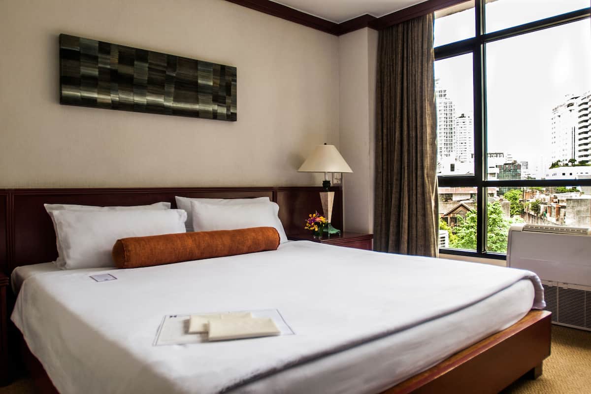 A hotel room with a large bed, a nightstand with a lamp, and a view of the city through the window.