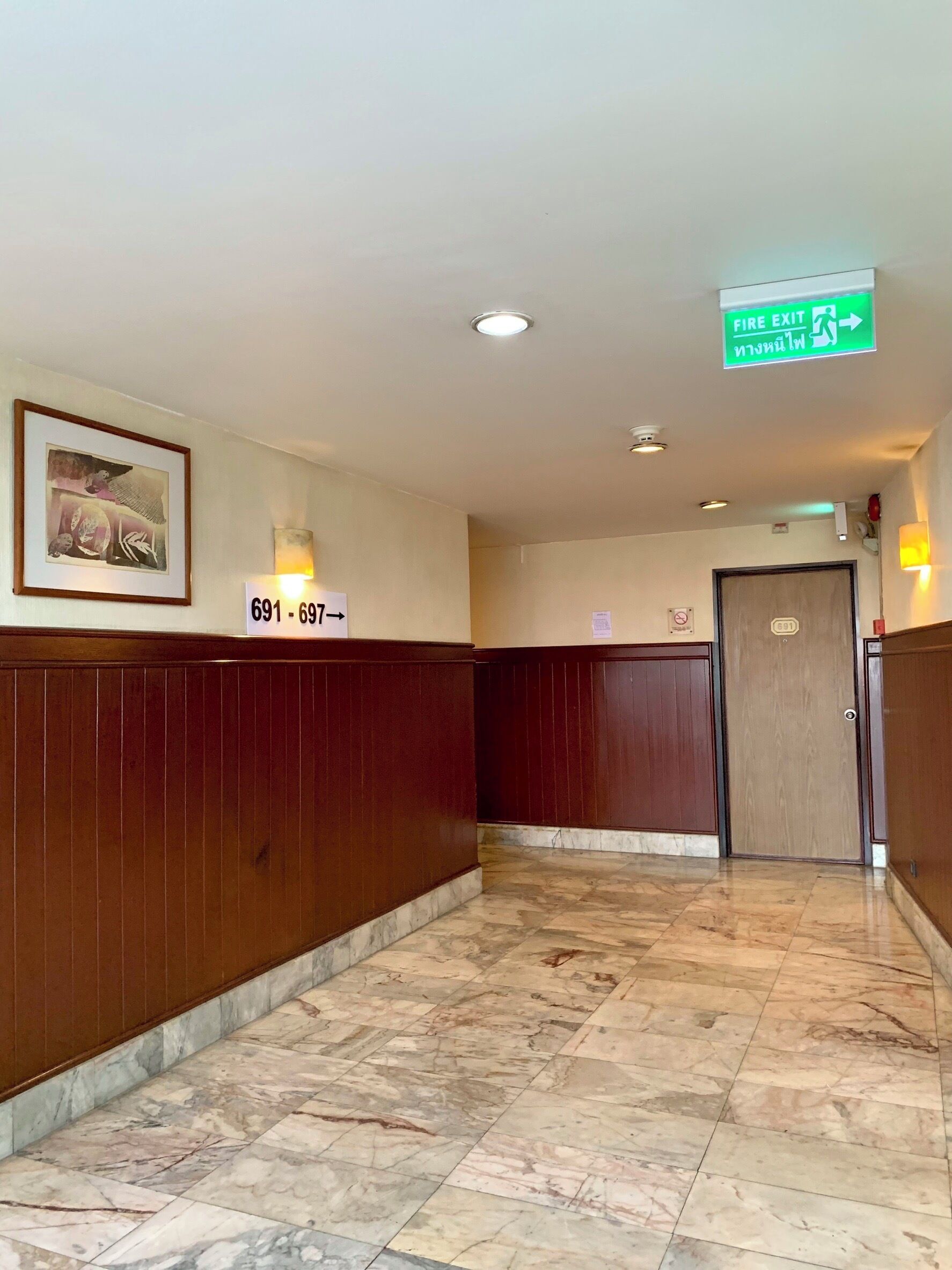 A hotel corridor with wooden paneling, a marble floor, a framed picture on the wall, and an exit sign.