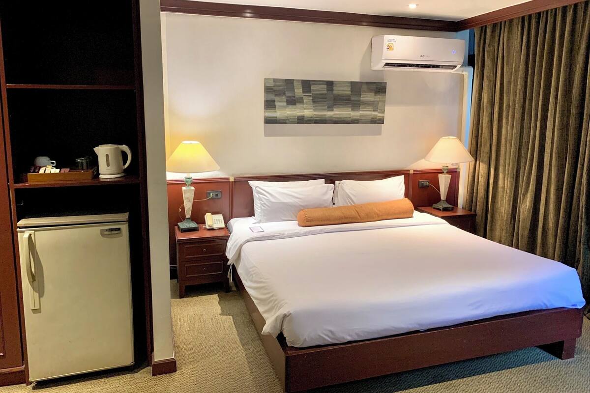 A hotel room with a bed, a small refrigerator, a bedside table with a lamp, a phone, and an air conditioning unit.