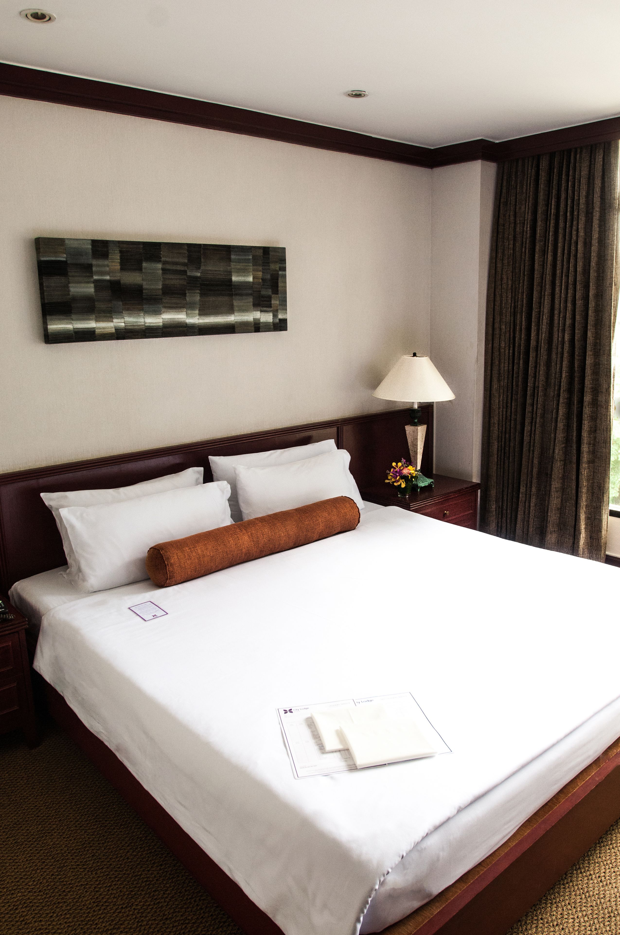 A hotel room with a neatly made bed, a nightstand with a lamp, a decorative wall piece, and a window with curtains.