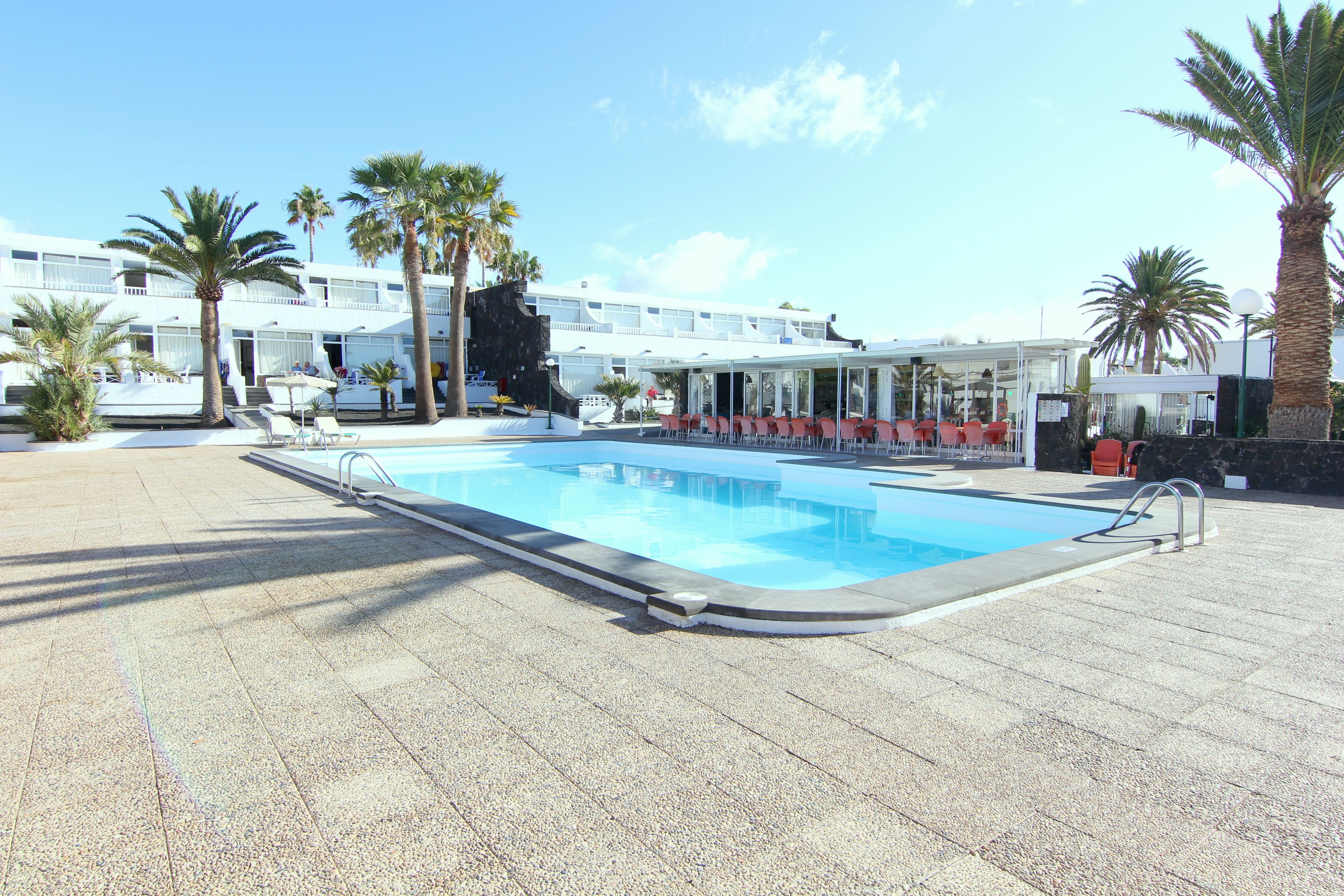 A swimming pool with a tiled deck, surrounded by palm trees and a building with balconies.