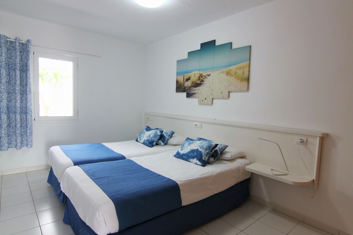 A bedroom with two beds, a window with curtains, a wall-mounted artwork, and a bedside table.