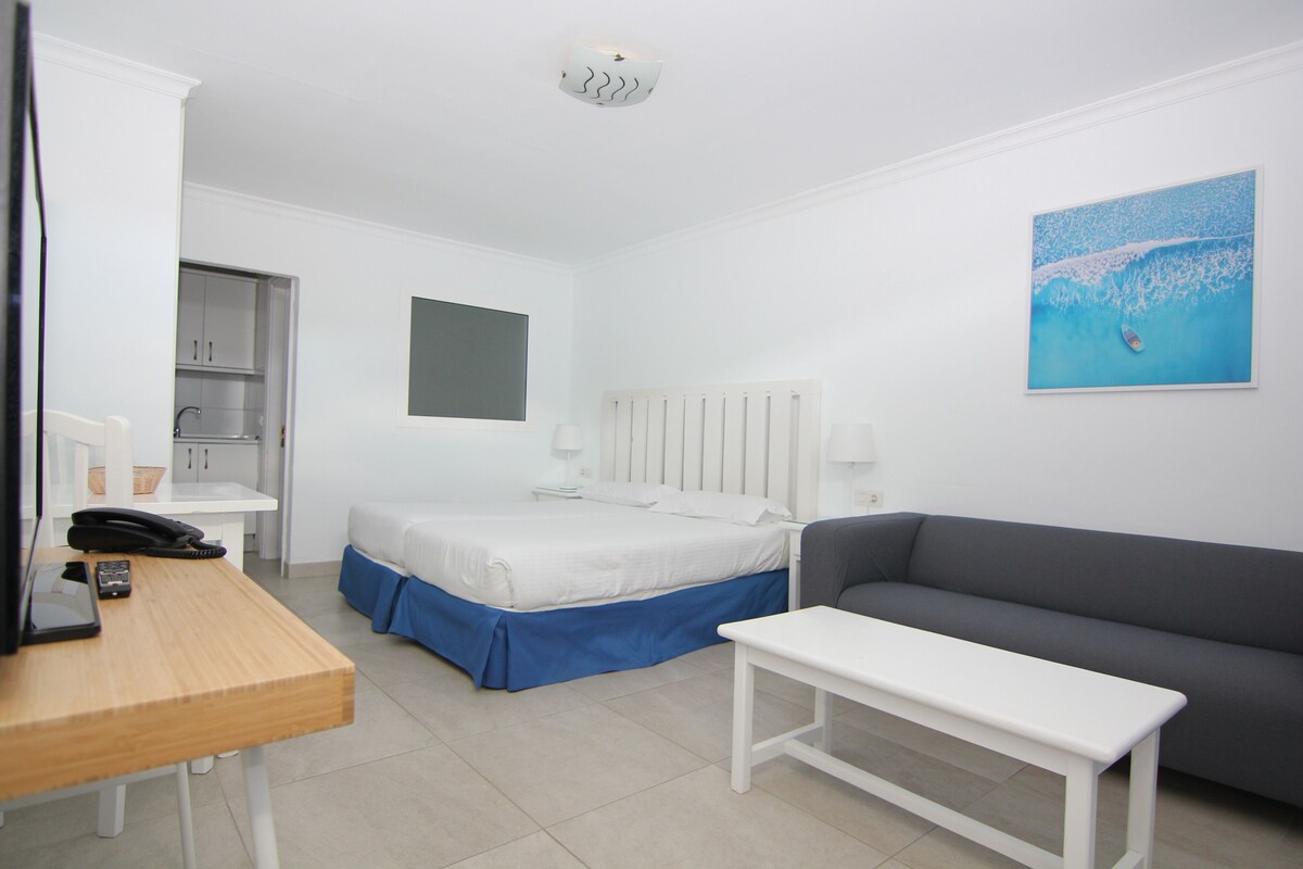 A hotel room with a bed, a grey sofa, a white bedside table, a desk with a telephone, and a wall-mounted painting.