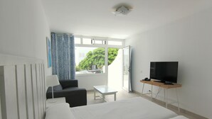 A hotel room with a bed, a desk, a chair, a TV, and a window with a view of greenery. - Arena Dorada - Apartments (Tías)