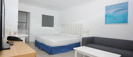 Habitación de hotel con cama, sofá gris, mesita de noche blanca, escritorio con teléfono y un cuadro colgado en la pared.