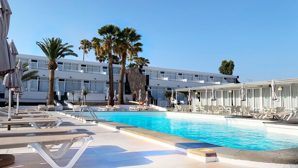 A pool area with lounge chairs and umbrellas, a clear blue pool, palm trees, and a modern building with multiple windows. - Arena Dorada - Apartments (Tías)