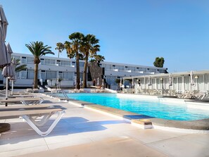 Un área de piscina con sillones de descanso y sombrillas, una piscina azul cristalina, palmeras y un edificio moderno con múltiples ventanas.
