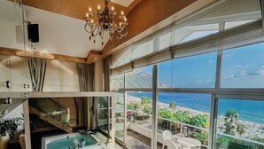 Een luxe badkamer met een ruime badkuip, een kroonluchter en uitzicht op een strand door grote ramen.