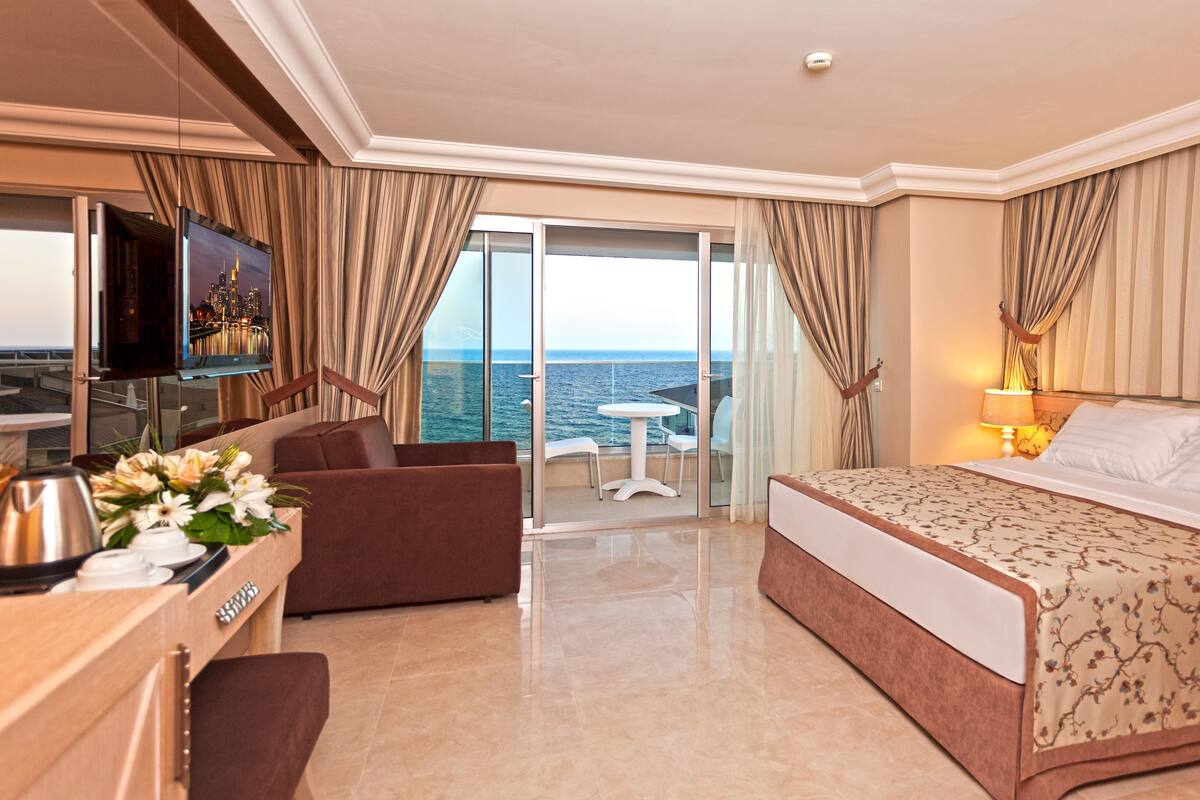 A hotel room with a bed, a desk with a coffee maker, a TV, and a balcony with a view of the sea.