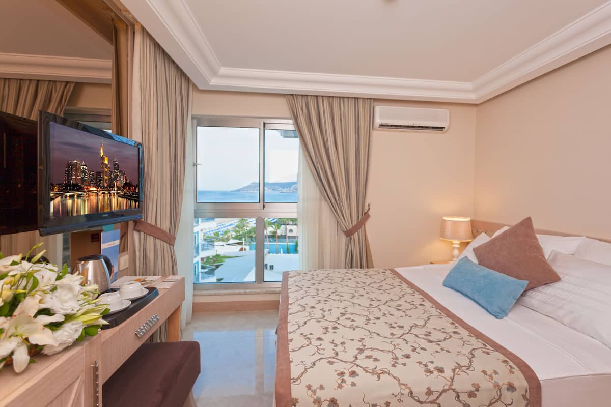 A hotel room with a bed, a desk with a TV, a view of the sea, and a balcony.