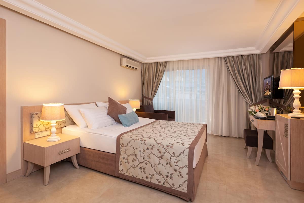 A hotel room with a bed, bedside tables, a desk, a TV, and a window with curtains.