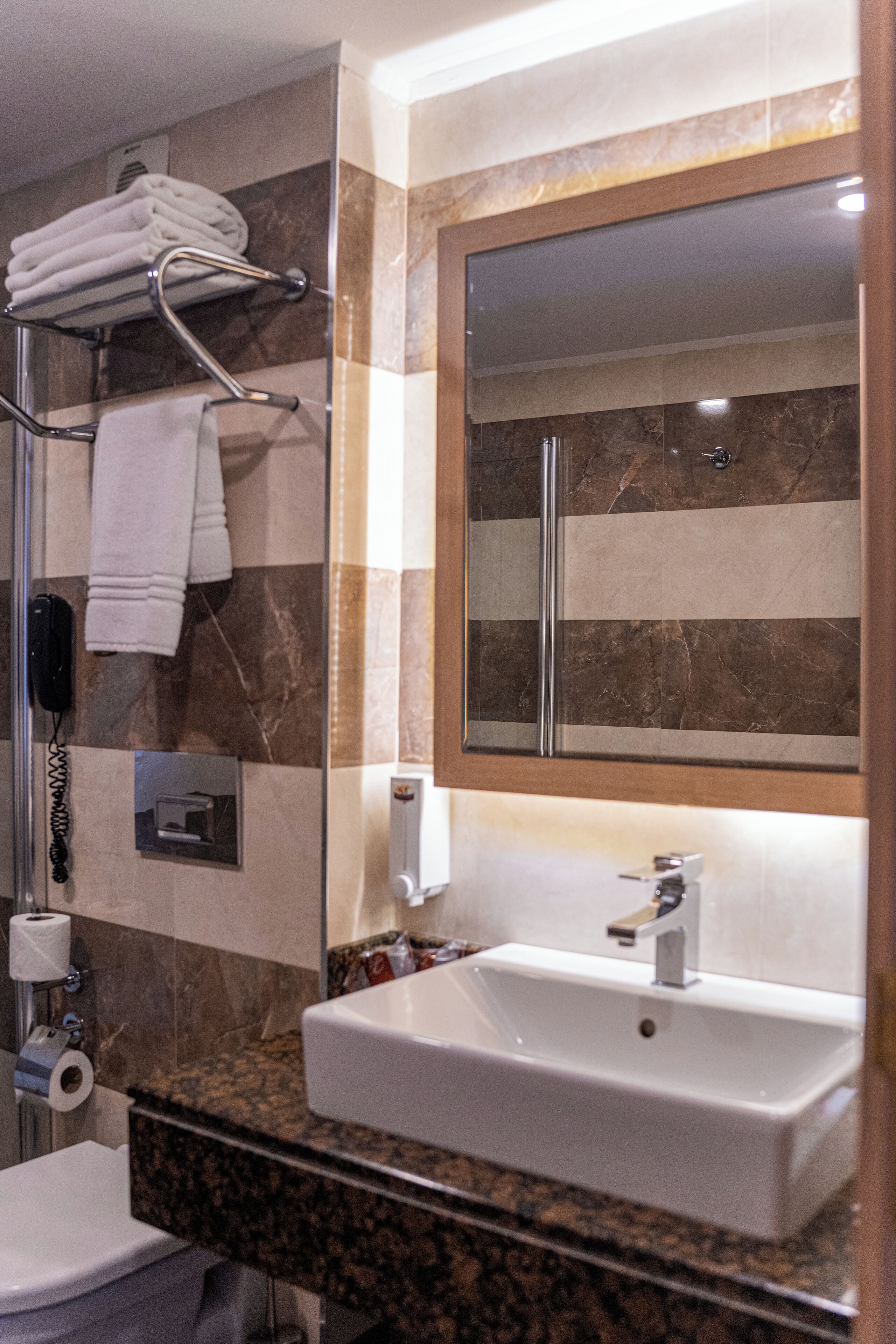 A modern bathroom with a marble-tiled wall, a large mirror, a white sink, and a towel rack with folded towels.